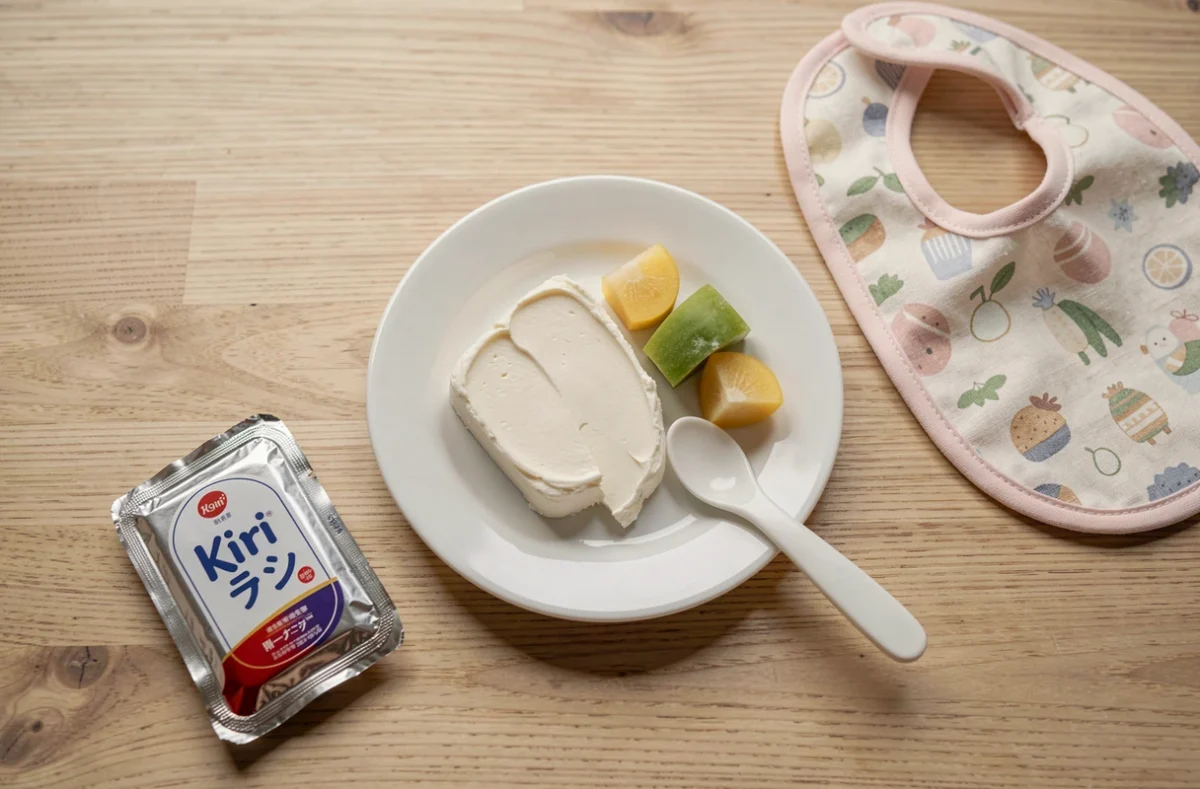 Présentation en flat-lay d'une portion de Kiri sur une assiette en céramique avec des légumes vapeur et une cuillère bébé sur une table en bois clair